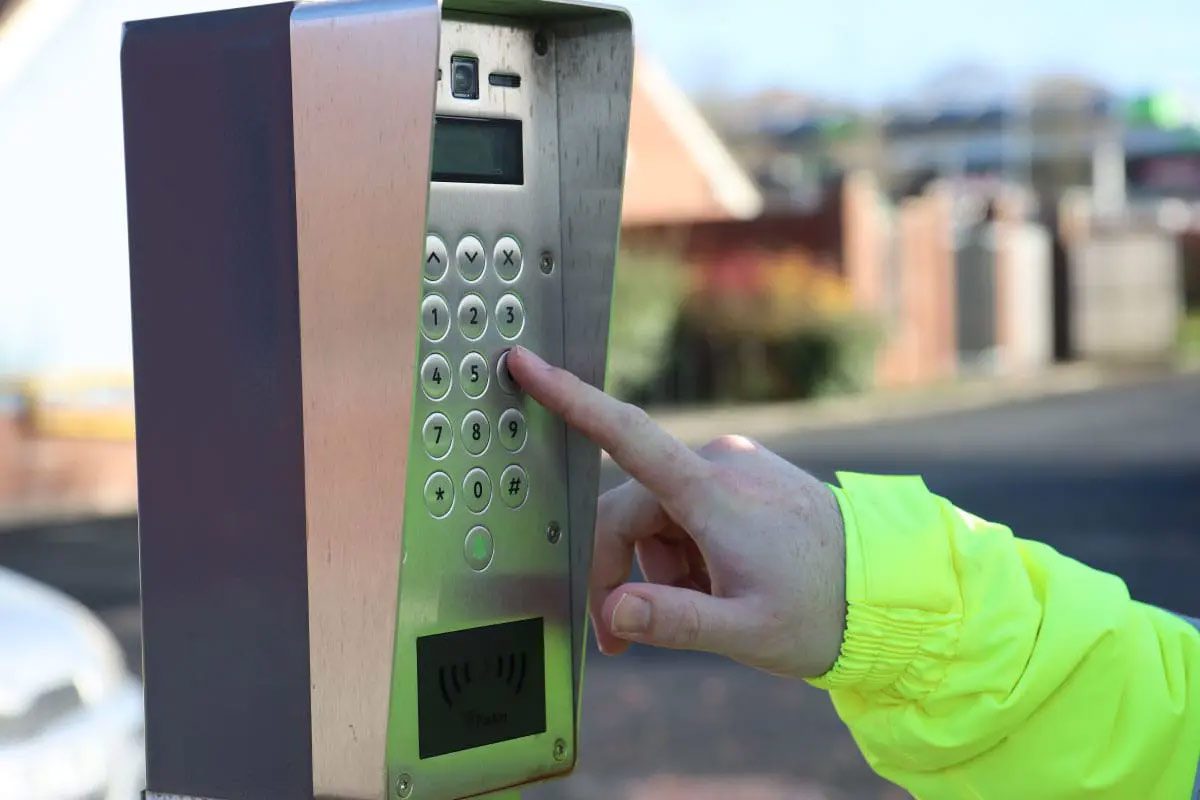 A close-up of a person in a bright yellow jacket pressing buttons on an outdoor metal intercom keypad