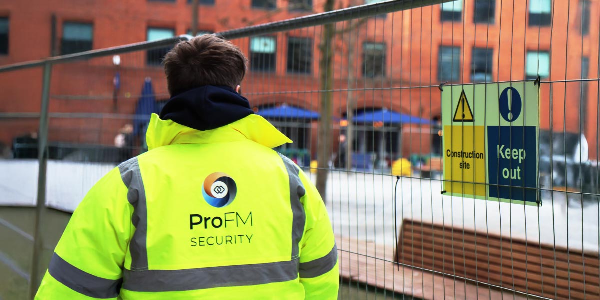 A ProFM security guard in a yellow high-visibility jacket, viewed from behind, looking through a construction fence