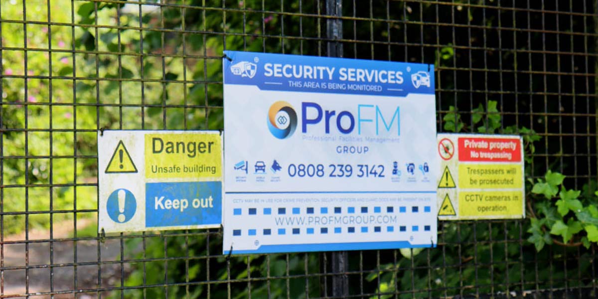 A metal wire fence displaying security warning signs, including a blue ProFM sign and a danger keep out sign