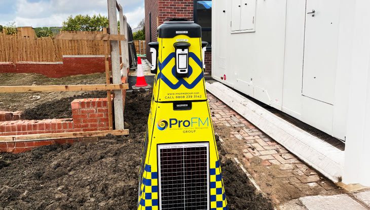 A yellow ProFM VPOD wireless security tower standing on muddy ground between a low brick wall and a white site cabin
