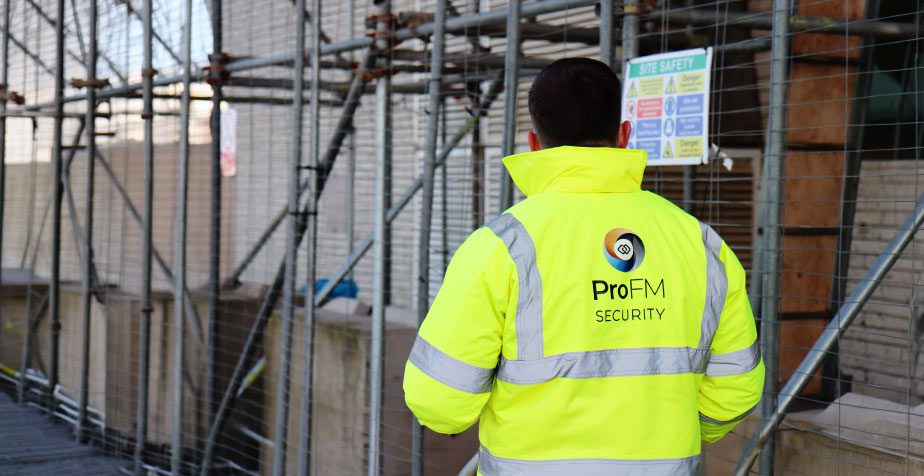 ProFM security officer inspecting perimeter fencing and access controls on an active construction site with scaffolding and site safety signage.