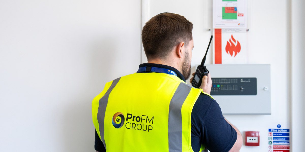 ProFM security officer using a two-way radio beside a fire alarm control panel while carrying out site security and facilities duties.